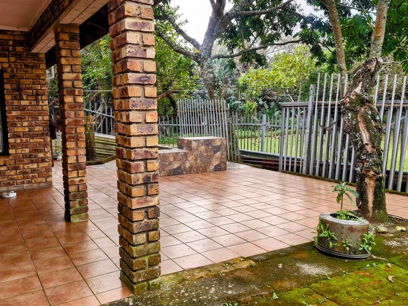 A covered outdoor patio featuring exposed brick pillars, tiled flooring, and built-in tiled seating, overlooking a garden area with metal perimeter fencing.