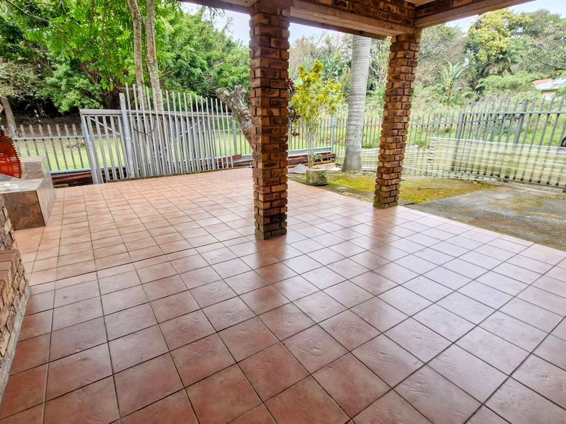 A covered outdoor patio area featuring brick support pillars and tiled flooring, overlooking a garden space with metal security fencing.