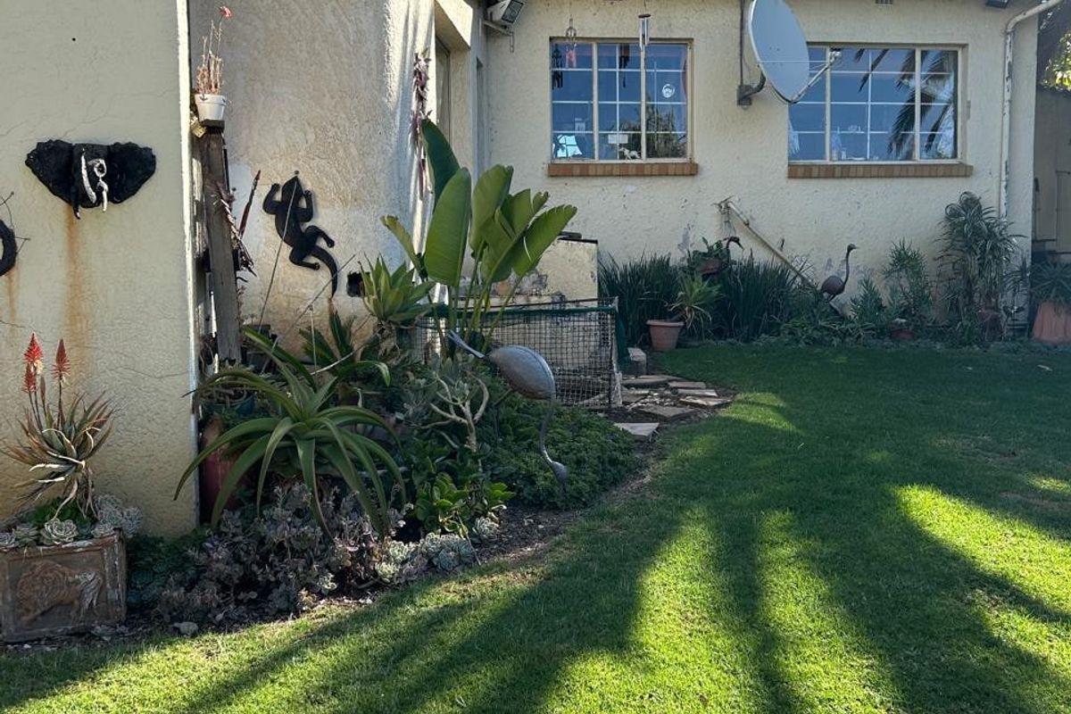 Rear view of the residential property featuring a lawn, established garden beds, and a tiled roof structure with exterior stucco finish.