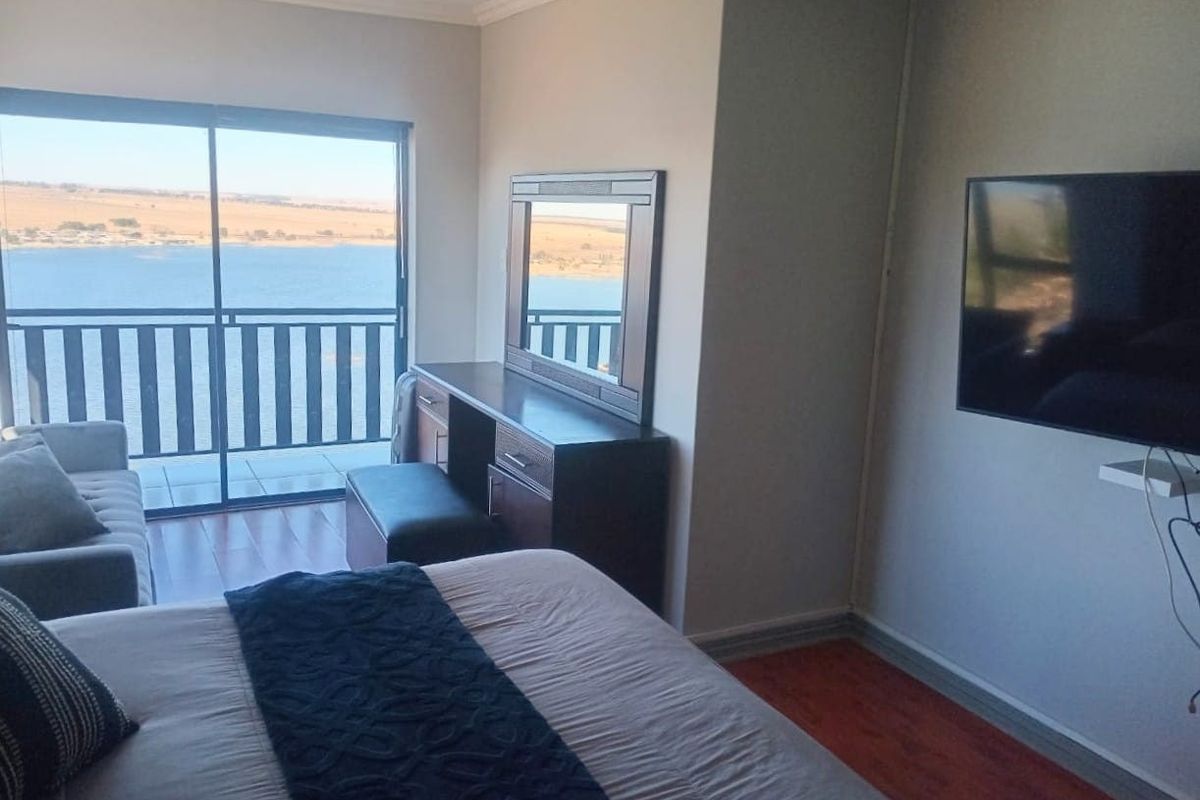 A well-lit bedroom featuring wood-look flooring, a wall-mounted television, and large sliding glass doors opening onto a balcony with scenic water views.