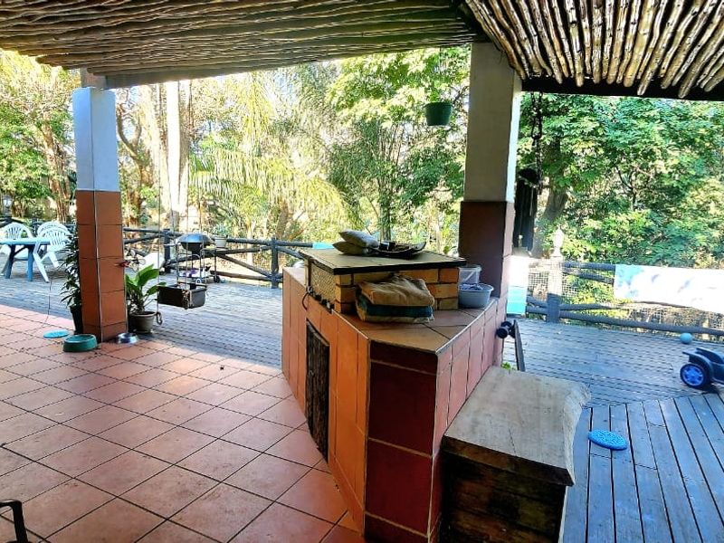 Covered outdoor patio featuring a built-in braai, tiled and timber deck flooring, and a rustic thatched roof overlooking a garden area.