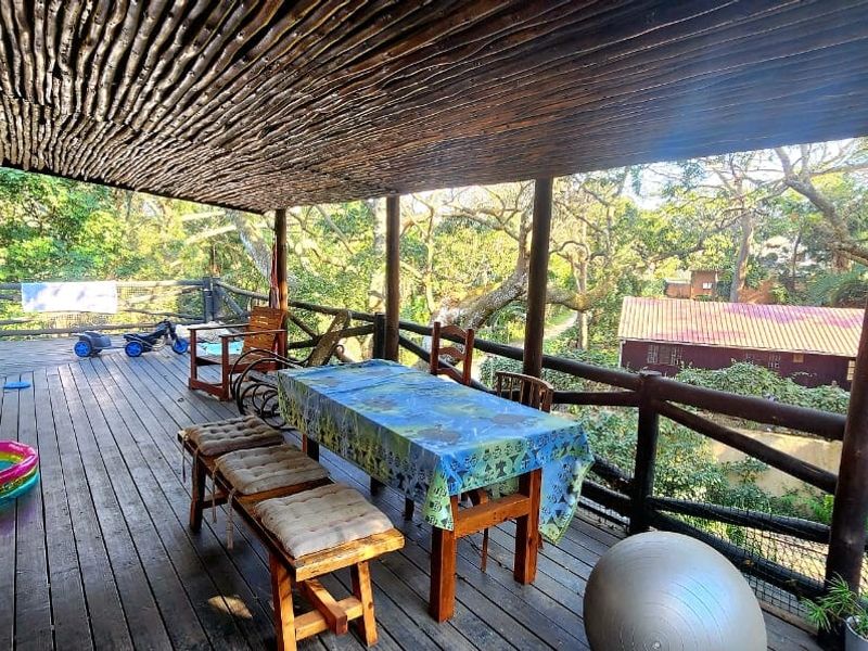 Spacious covered timber deck featuring a rustic pole roof structure, timber balustrades, and a built-in brick braai, overlooking a lush garden setting.