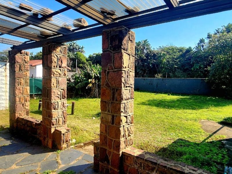 A covered outdoor patio featuring rustic stone pillars and flagstone flooring, overlooking a spacious backyard with established greenery.