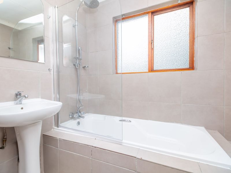 A functional bathroom featuring a pedestal basin, shower-over-bath combination with a glass screen, and neutral wall tiling.