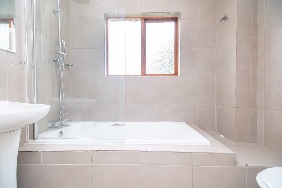 A functional bathroom featuring a built-in bathtub with a glass shower screen, pedestal basin, and neutral wall tiling.