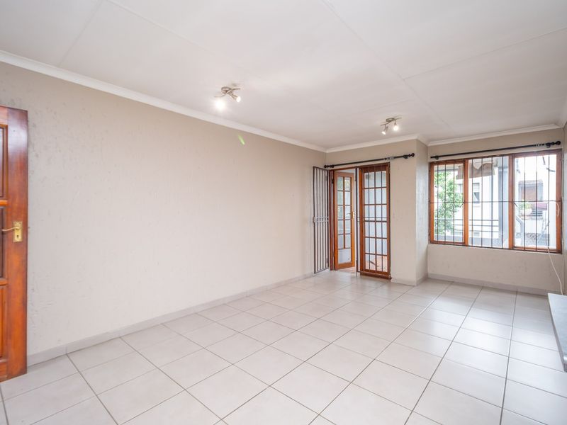 A bright, empty living area featuring neutral tiled flooring, wooden door access, and large windows with security bars, offering a clean canvas for styling.