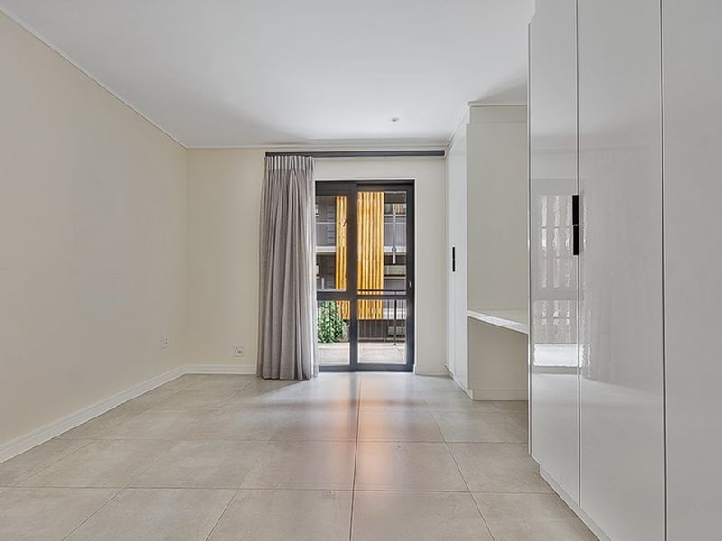 A bright, modern bedroom featuring neutral tiled flooring, ample built-in cupboard space, a built-in desk, and direct access to a balcony through sliding doors.