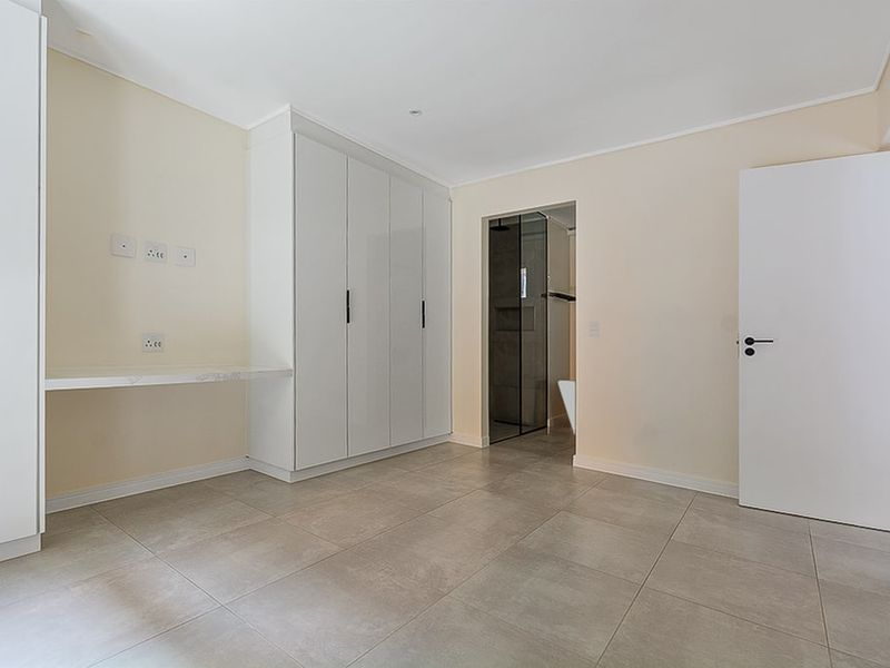 A clean, modern bedroom featuring neutral tiled flooring, ample built-in cupboard space, a floating desk, and direct access to an en-suite bathroom.
