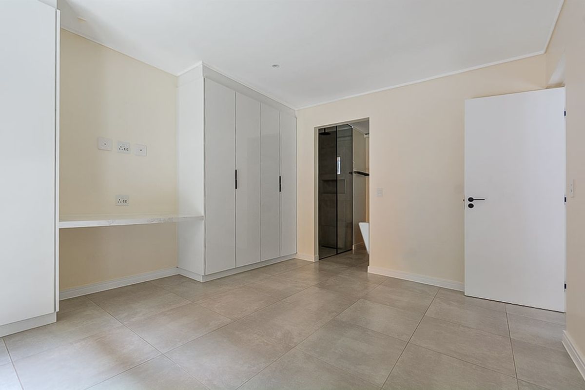 A clean, modern bedroom featuring neutral tiled flooring, ample built-in cupboard space, a floating desk, and direct access to an en-suite bathroom.