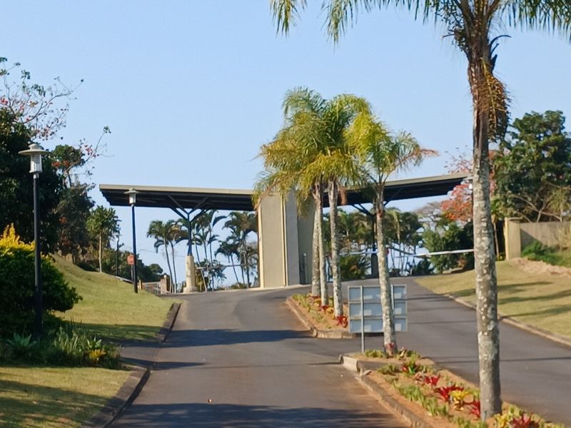 A well-maintained entrance to a residential estate featuring a security gatehouse, boom gate, paved driveway, and landscaped gardens.