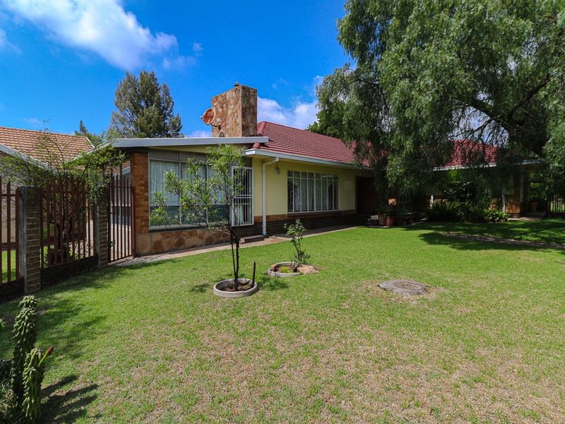 Rear view of the house featuring a stone chimney, tiled roof, and a spacious garden with established trees and secure metal fencing.