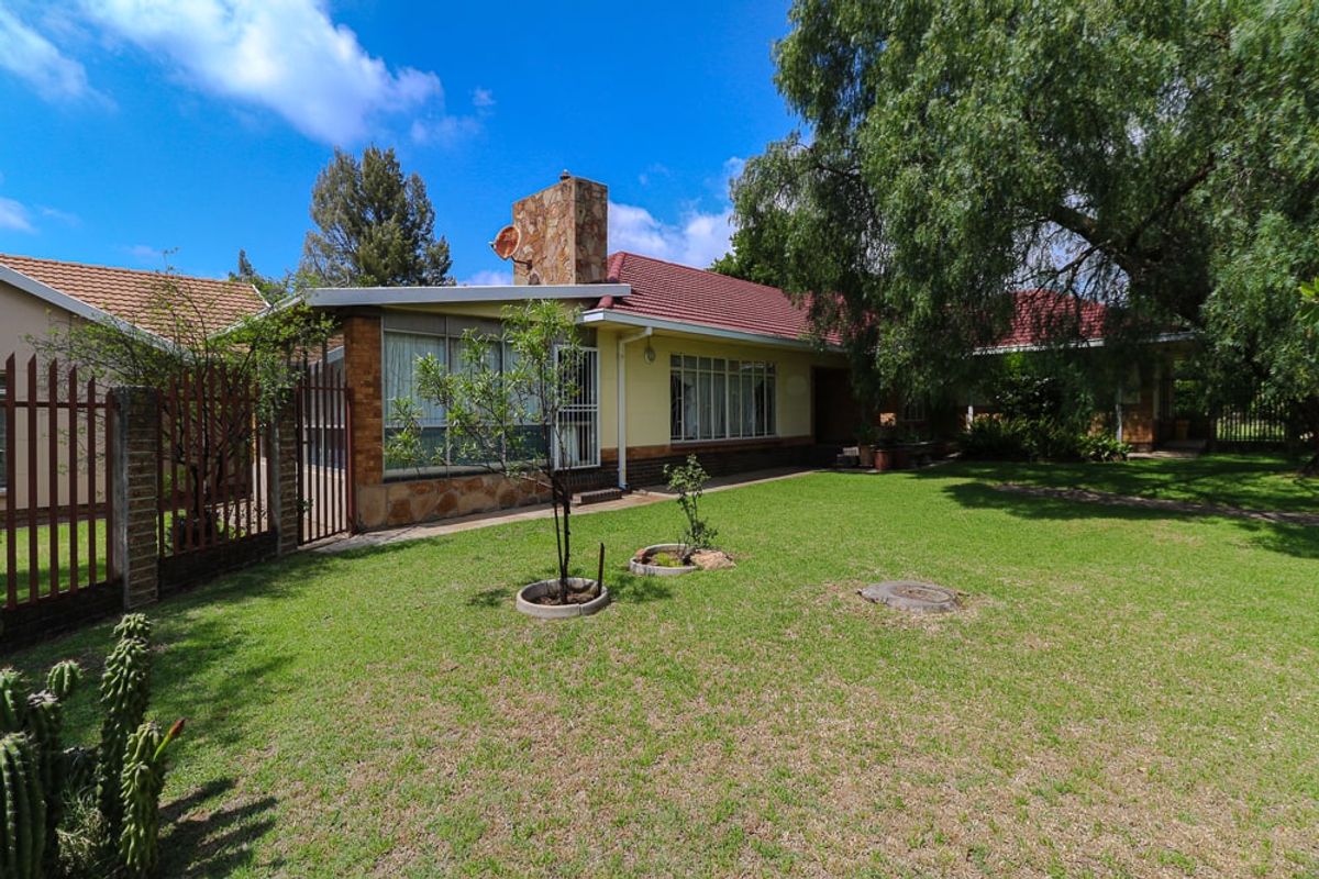 Rear view of the house featuring a stone chimney, tiled roof, and a spacious garden with established trees and secure metal fencing.