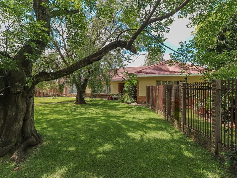Front view of a residential house featuring a well-maintained lawn, mature trees, and secure metal perimeter fencing with a tiled roof structure.