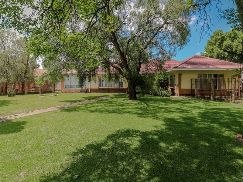 A spacious rear garden featuring a well-maintained lawn, mature trees, a paved walkway, and a view of the home's rear brick facade.