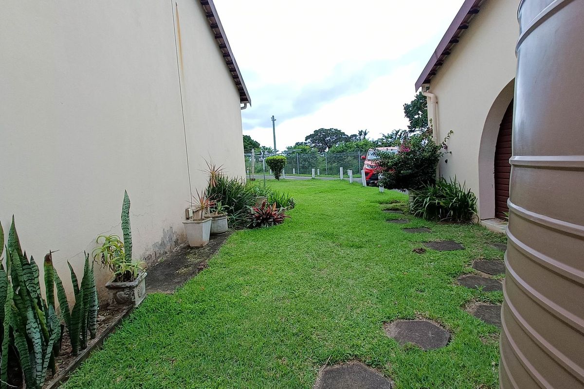 A side garden area between two townhouse structures featuring a lawn, stepping stone path, and a large water tank.