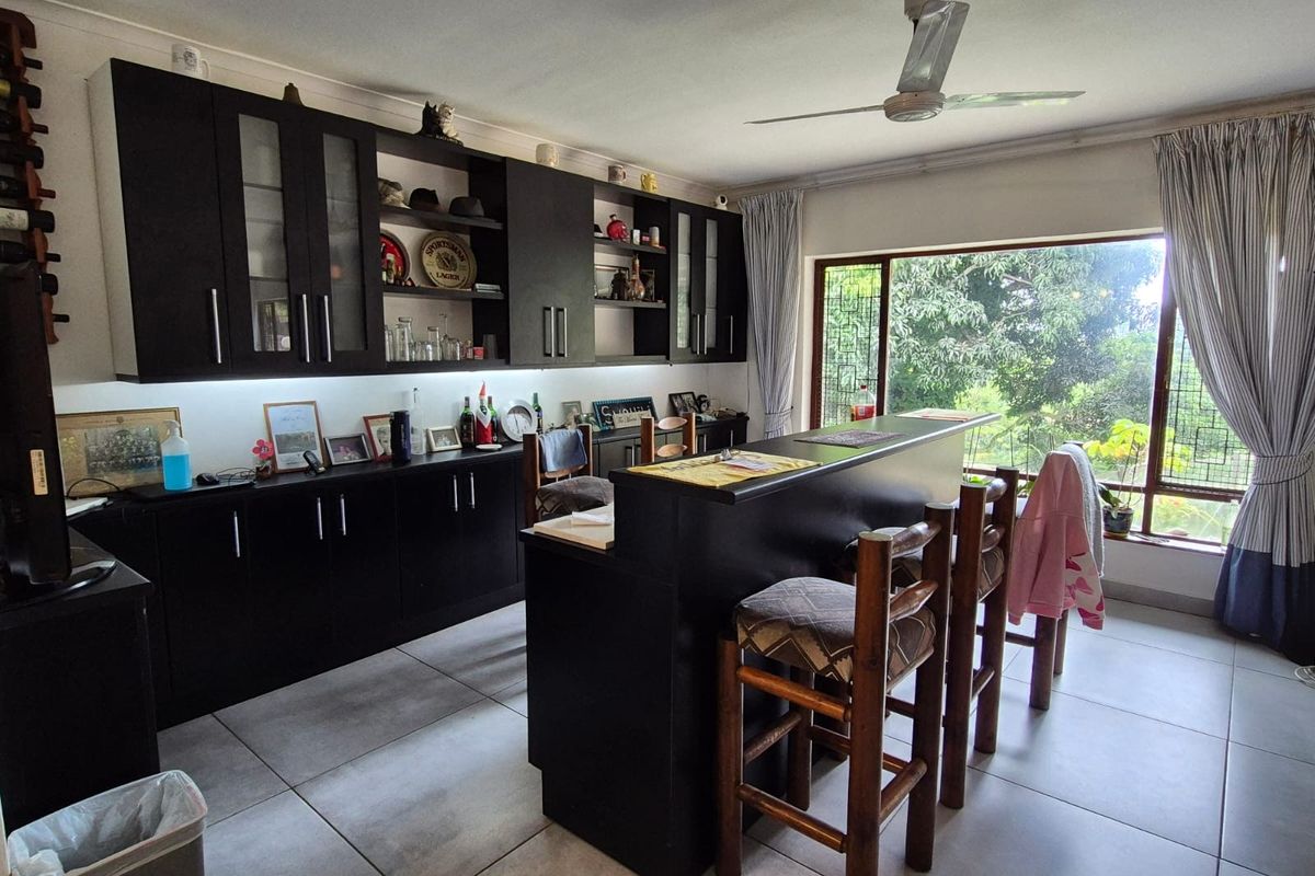 A functional kitchen featuring dark built-in cabinetry, a central breakfast bar, tiled flooring, and a ceiling fan with large windows overlooking the garden.