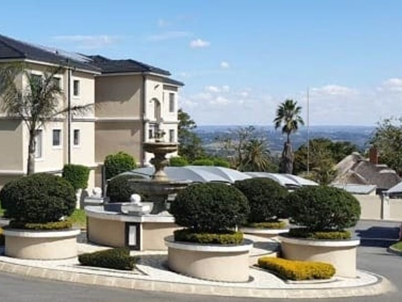A well-maintained apartment complex entrance featuring a central stone fountain, manicured circular planters, and paved driveway with scenic elevated views.