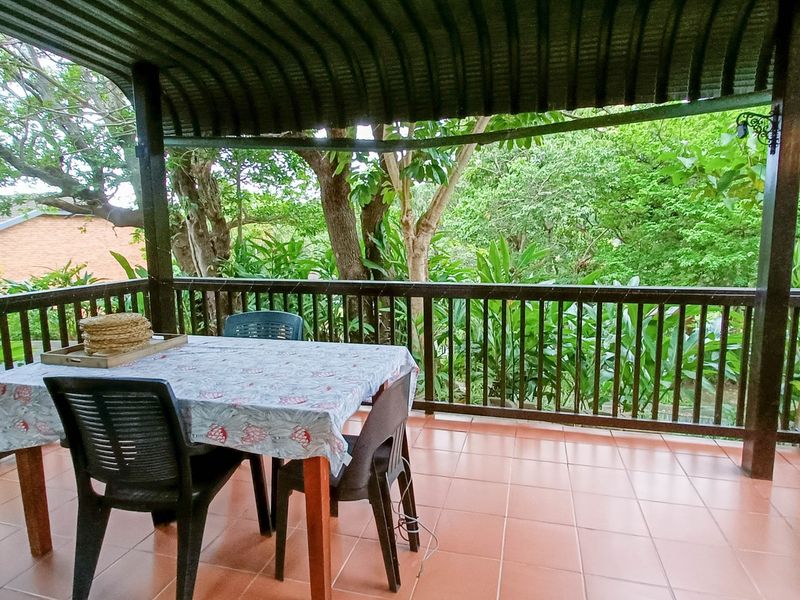 A covered outdoor patio featuring tiled flooring, a timber balustrade, and a corrugated metal roof, overlooking a lush garden setting.