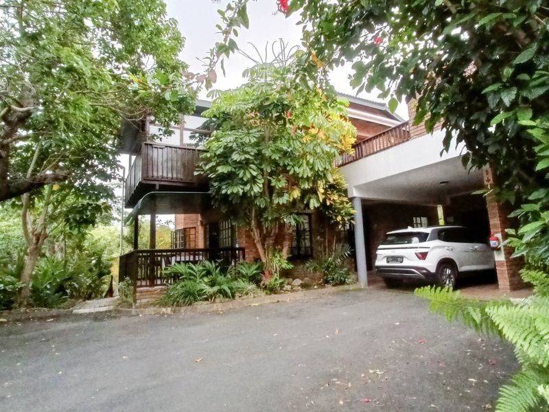Front exterior view of a brick apartment building featuring a timber balcony, covered carport, and mature surrounding garden.
