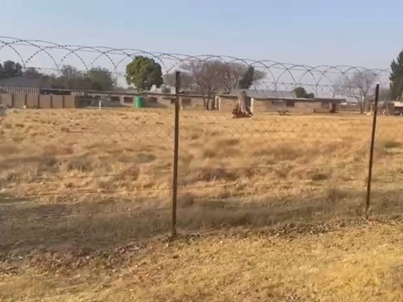 A view of a vacant residential plot featuring dry grass, enclosed by a perimeter wall and topped with security razor wire fencing.