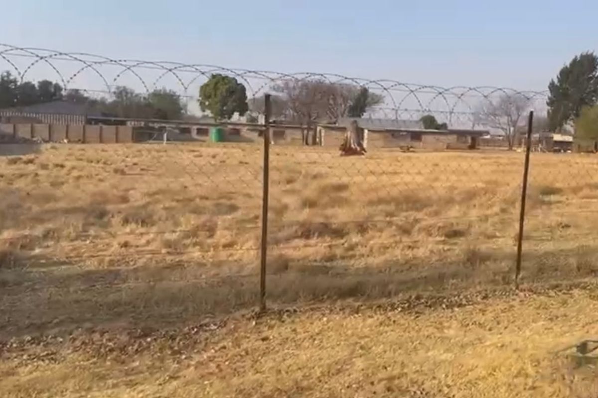 A view of a vacant residential plot featuring dry grass, enclosed by a perimeter wall and topped with security razor wire fencing.