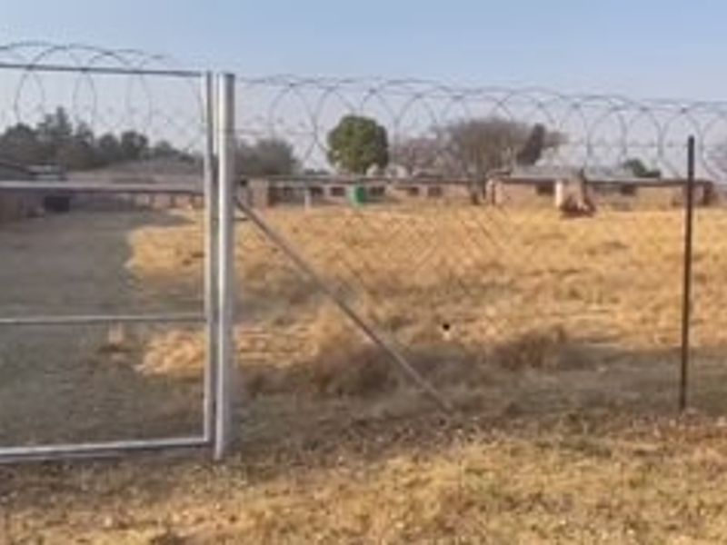 A vacant residential plot enclosed by chain-link fencing topped with razor wire, featuring a metal gate entrance.