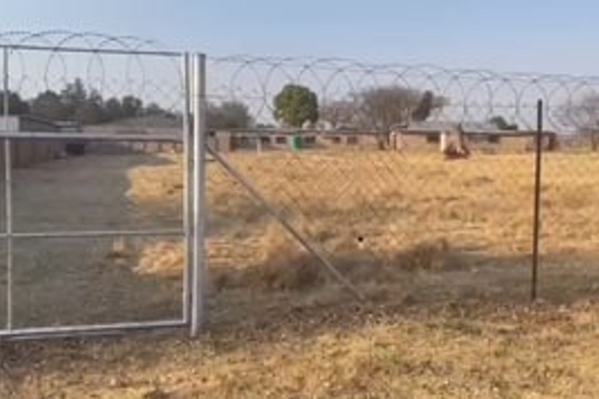 A vacant residential plot enclosed by chain-link fencing topped with razor wire, featuring a metal gate entrance.