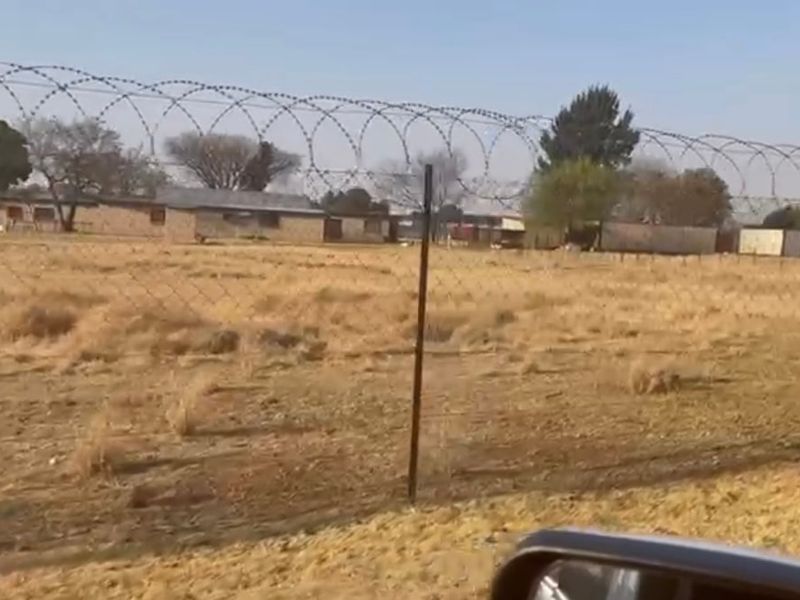 A view of a vacant residential plot enclosed by a chain link fence topped with razor wire, showing dry grass and surrounding structures.
