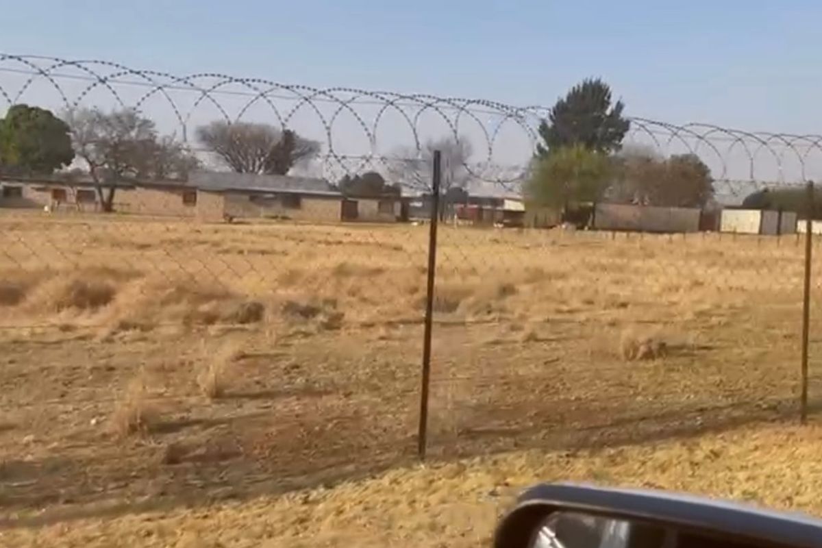 A view of a vacant residential plot enclosed by a chain link fence topped with razor wire, showing dry grass and surrounding structures.