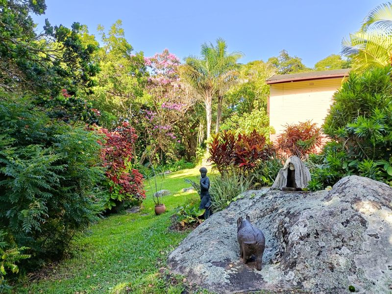 A lush, well-maintained garden featuring mature trees, vibrant foliage, and a prominent natural rock formation in a peaceful outdoor setting.
