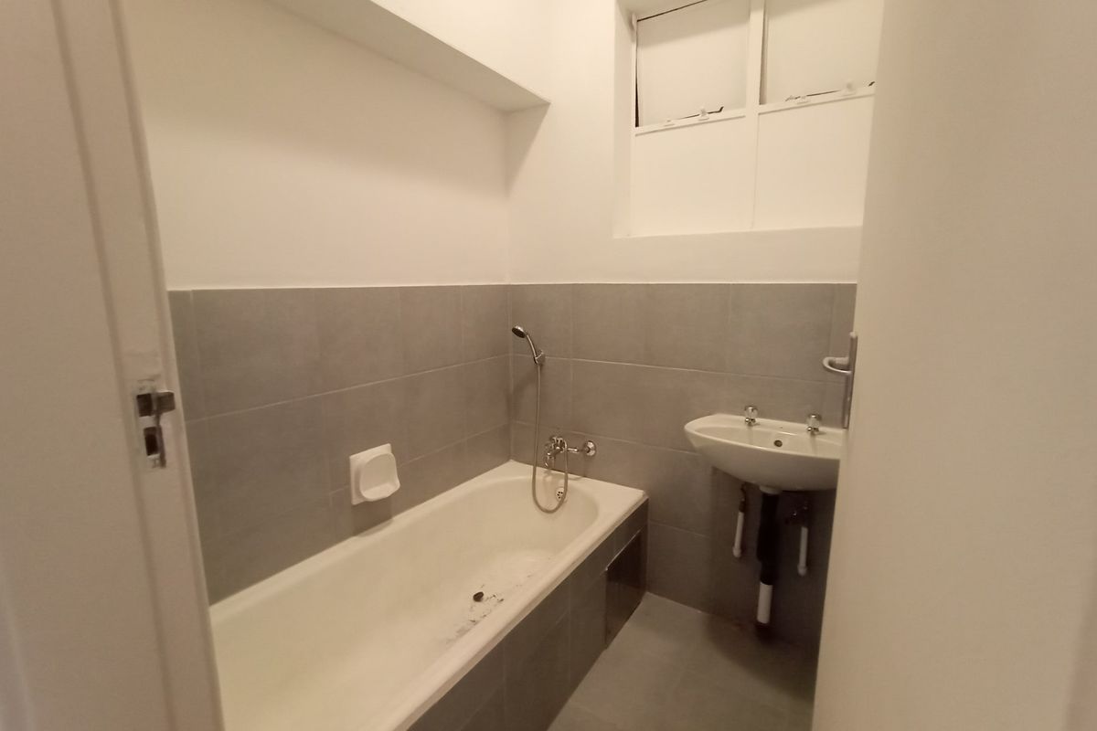 A functional bathroom featuring a bathtub with handheld shower mixer, wall-mounted basin, and neutral grey wall and floor tiling.