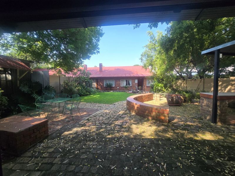 A spacious rear garden featuring brick paving, a curved retaining wall, a built-in braai area, and a covered patio overlooking the lawn.