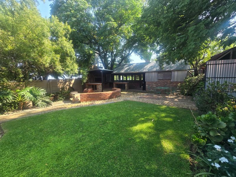 A spacious rear garden featuring a brick-built outdoor braai area, paved patio space, and established trees, offering a private outdoor entertainment setting.