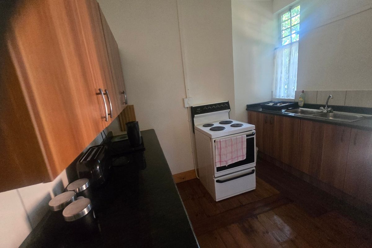 A functional kitchen featuring wooden cabinetry, black countertops, a freestanding electric stove, and a stainless steel double sink with timber flooring.