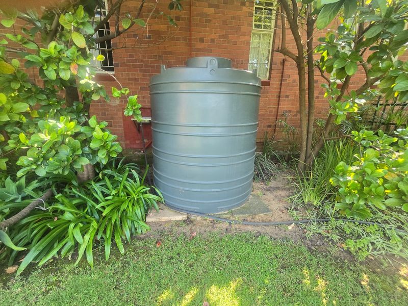 A grey plastic water storage tank positioned against the rear brick wall of the house, surrounded by garden landscaping.