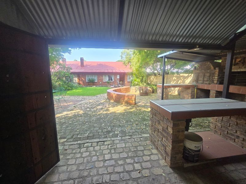 Covered outdoor patio area featuring a built-in brick braai, brick counter, and paved flooring, overlooking a rear garden and main house structure.