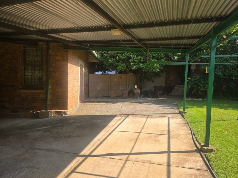 A covered outdoor patio area featuring a corrugated metal roof, steel support structure, and concrete paving, adjacent to the main brick building.
