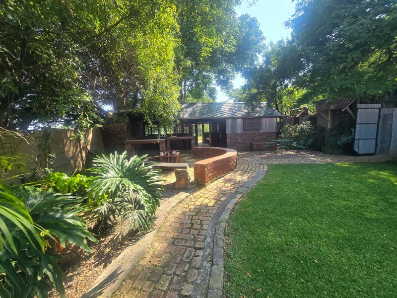 A spacious rear garden featuring a brick-paved pathway, a curved brick retaining wall, and a built-in outdoor braai area under a corrugated metal roof.