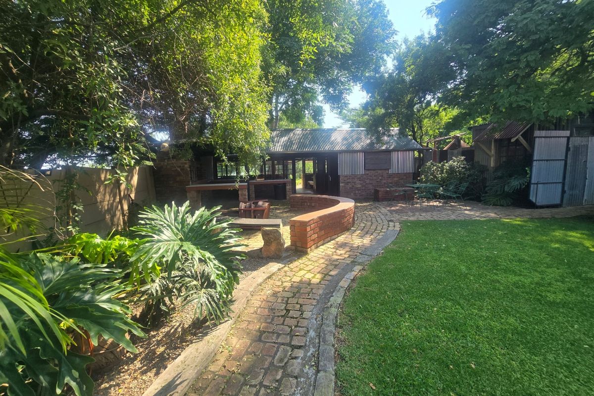 A spacious rear garden featuring a brick-paved pathway, a curved brick retaining wall, and a built-in outdoor braai area under a corrugated metal roof.