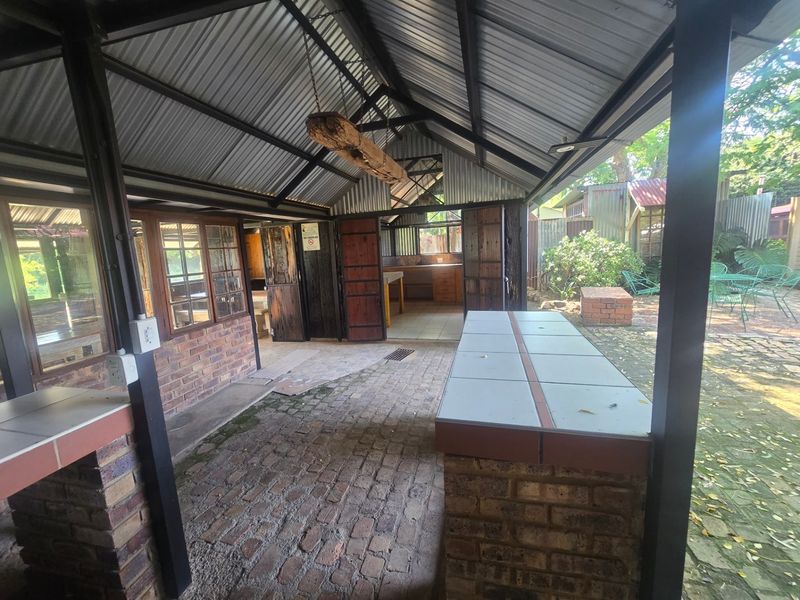 Covered outdoor patio area featuring a brick-built counter with tiled top, corrugated metal roofing, and exposed steel structure, overlooking a garden space.