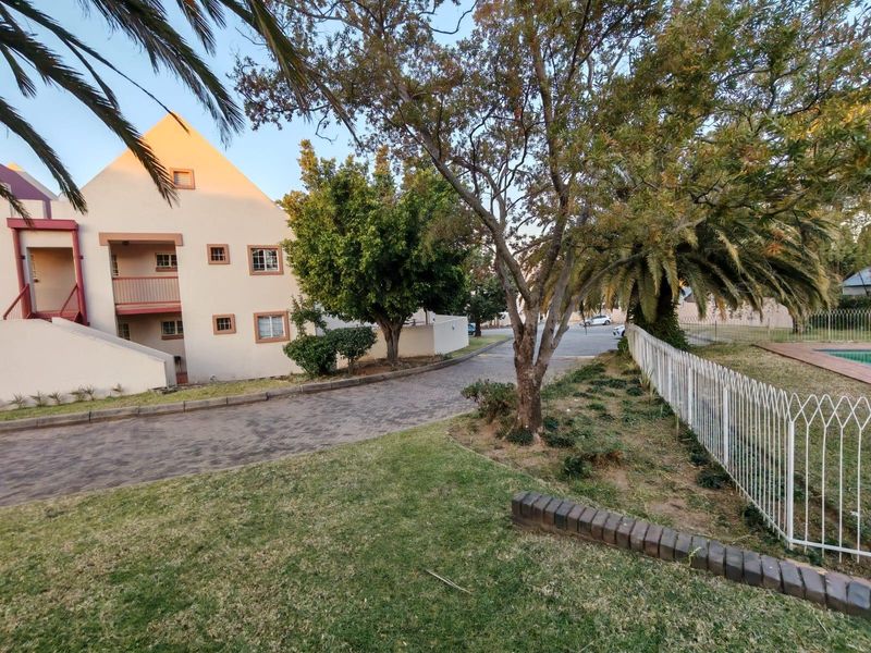 Exterior view of an apartment complex featuring a paved driveway, landscaped garden areas, metal perimeter fencing, and building balconies.