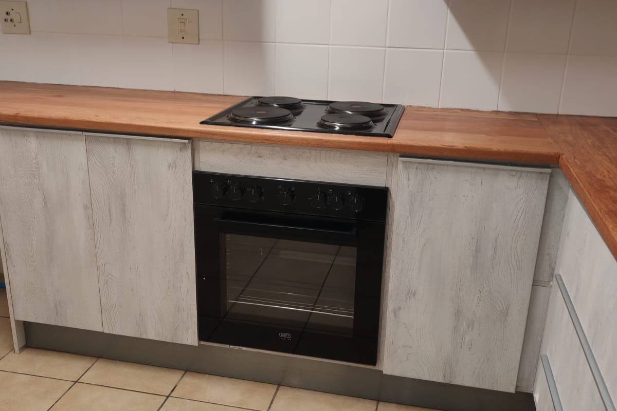 A clean kitchen area featuring light wood-grain cabinetry, a warm wooden countertop, a built-in oven with an electric hob, and neutral ceramic floor tiles.