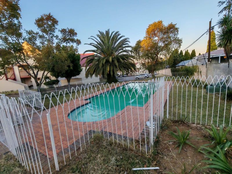 Outdoor swimming pool area featuring brick paving, secure metal fencing, and established garden surroundings within an apartment complex.