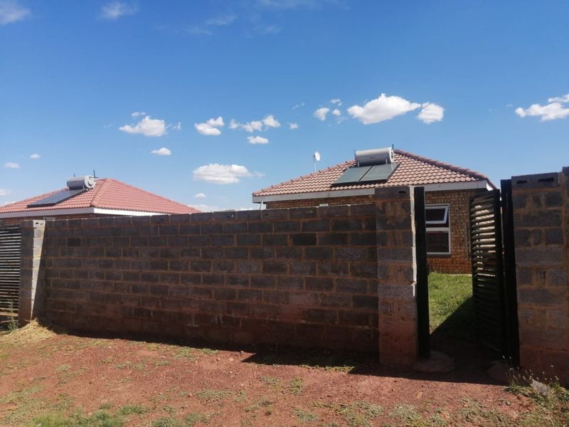 Front view of a residential property featuring a brick facade, tiled roof with solar water heating system, and a concrete block boundary wall.