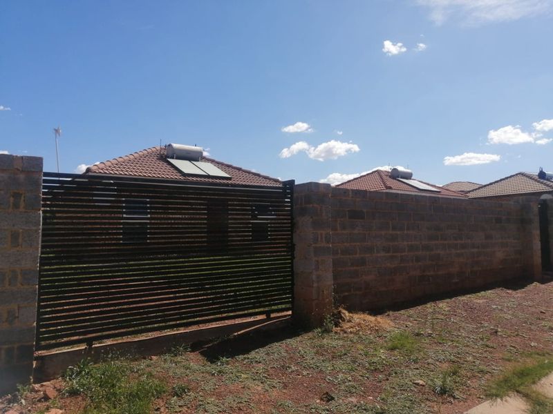Front view of a residential property featuring a brick boundary wall, slatted metal gate, and tiled roof with an installed solar water heating system.