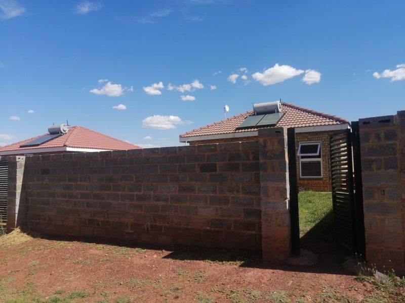Front view of a residential property featuring a brick facade, tiled roof with solar water heater, and a concrete block boundary wall with gate.