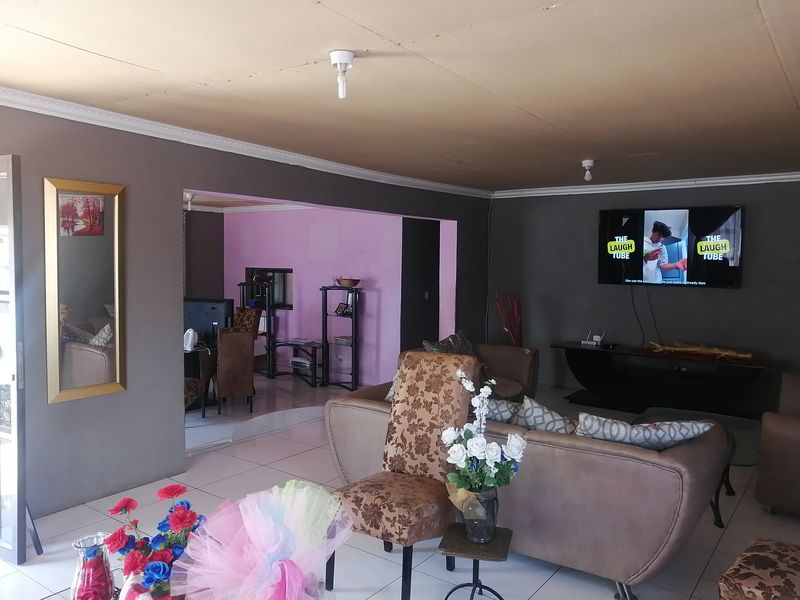 A living room featuring tiled flooring, cove cornicing, and a wall-mounted television, with an adjacent dining area visible through the opening.