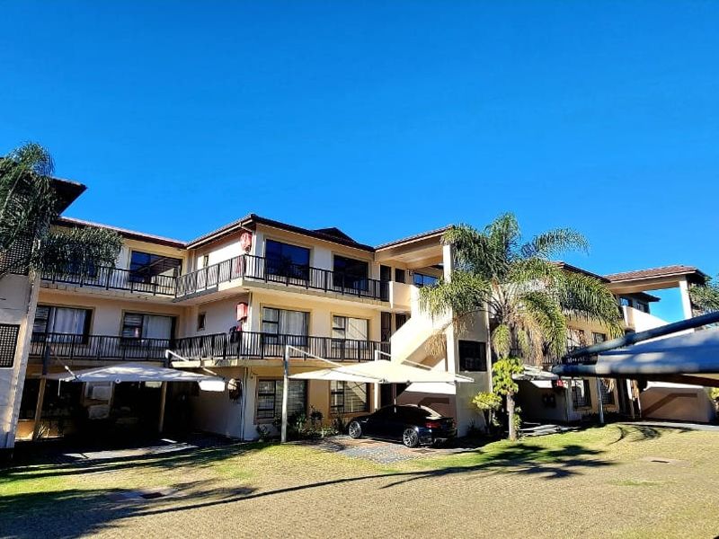 A multi-story apartment complex featuring balconies with railings, tiled roofing, and covered parking bays within a landscaped residential setting.