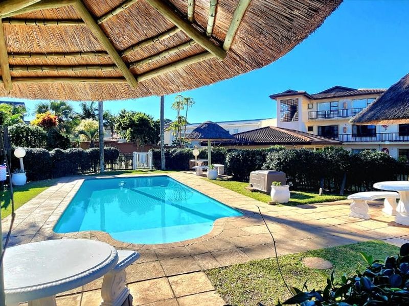 Outdoor pool area featuring a swimming pool, paved surround, and thatched umbrellas within a landscaped complex setting.