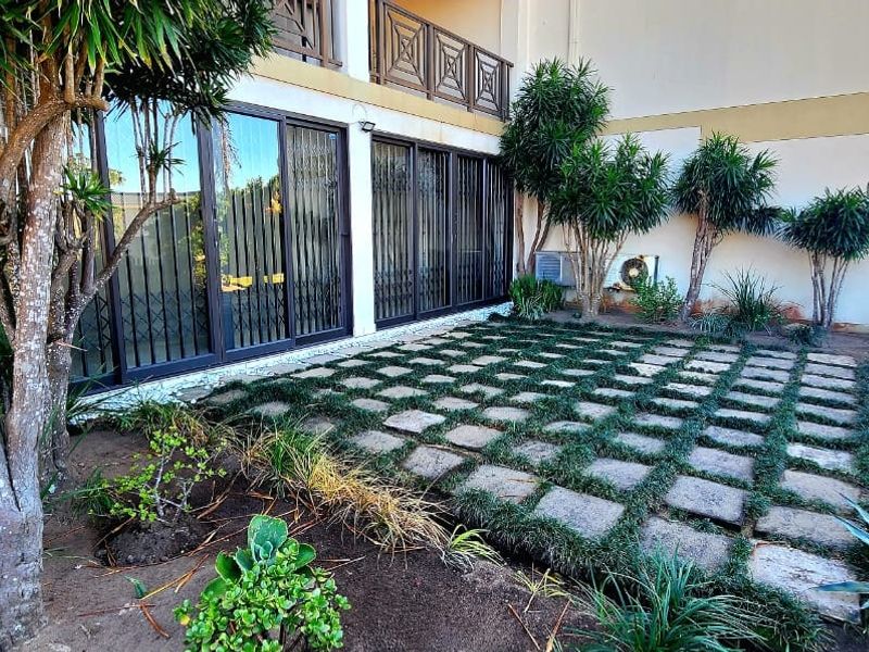 A rear exterior view of the apartment featuring a paved patio area, sliding glass doors with security gates, and an upper-level balcony.